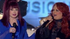 Naomi Judd is wearing a blue jacket and her hair is half up as holds a microphone with her right hand. Wynonna Judd is wearing her hair down and a black shit as she holds a microphone in her right hand. They are singing a duet.