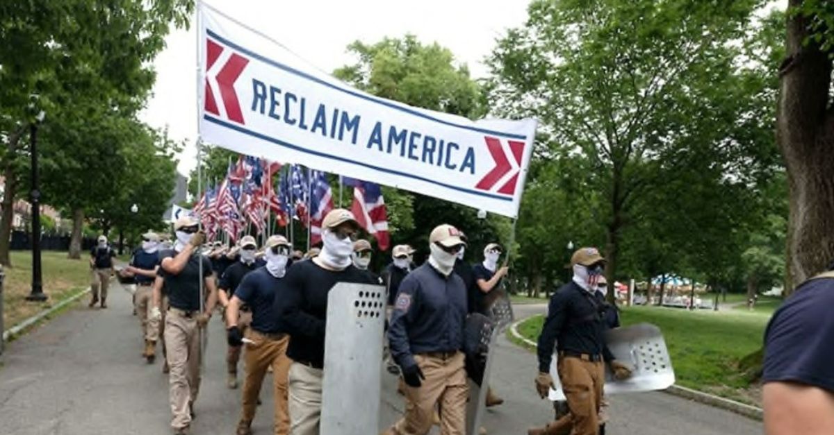 Members of Patriot Front march down a trail carrying a large banner that states "Reclaim America." They are masked and wear similar clothing, including dark colored shirts and khaki pants. Their faces are obscured with sunglasses, gaiters and masks. 